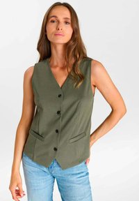 Femme aux longs cheveux bruns portant un gilet sans manches vert olive boutonné et un jean bleu clair, debout devant un fond blanc uni.