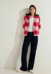 Red and white plaid jacket with a black hood, worn over a white shirt. Dark blue wide-leg jeans and white sneakers complete the outfit.