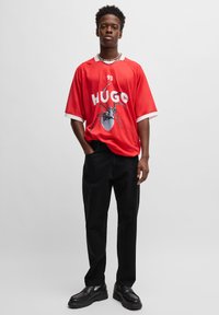 Rød oversized t-skjorte med hvite ermer, med "HUGO" og et edderkoppgrafikk. Kombinert med svarte bukser med rett ben og kraftige svarte sko.
