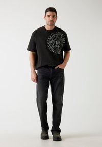 Zwarte graphic t-shirt met grote logo-print, gecombineerd met donkerblauwe jeans en grijze schoenen. Eenvoudige, casual urban outfit.