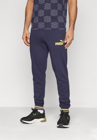 Pantalones de chándal navy con un logo dorado de Puma, detalles en los puños acanalados, combinados con zapatillas deportivas negras y verdes, que cuentan con una camiseta de patrón geométrico.