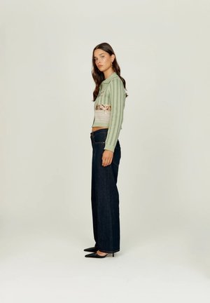 Naine seisab külje pealt, seljas rohelist värvi triibuline lühike kampsun, millel on heegeldatud detailid, ning tumedad laia säärega teksapüksid ja mustad terava ninaga kontsakingad.