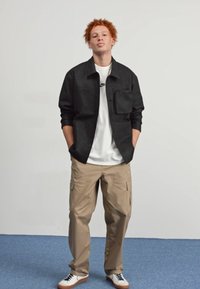 Sort button-up jakke med en brystlomme, hvid t-shirt, beige cargo-bukser, og hvide sneakers med brune accenter. Neutral baggrund.
