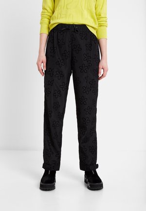 Osoba oblečená v černých kalhotách s květinovými vzory, žlutém svetru a černých platformových botách stojící před bílým pozadím.