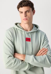 Joven con cabello castaño que lleva puesta una sudadera verde claro con el logotipo rojo "HUGO", brazos cruzados, expresión neutral, fondo liso claro.