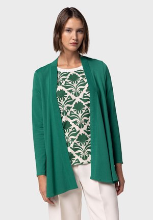 Cardigan vert avec un devant ouvert, manches longues, associé à un haut à motifs présentant des designs floraux verts sur un fond clair.