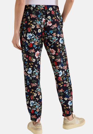 Vue de dos d'une personne portant un pantalon noir à imprimé floral et des baskets beiges, debout avec la main droite dans la poche sur fond blanc.