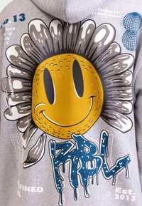 Szara bluza z kapturem z dużym wzorem uśmiechniętego słońca o teksturowanej żółtej twarzy, otoczonego białymi i czarnymi płatkami, oraz niebieskim tekstem w stylu dripping poniżej.