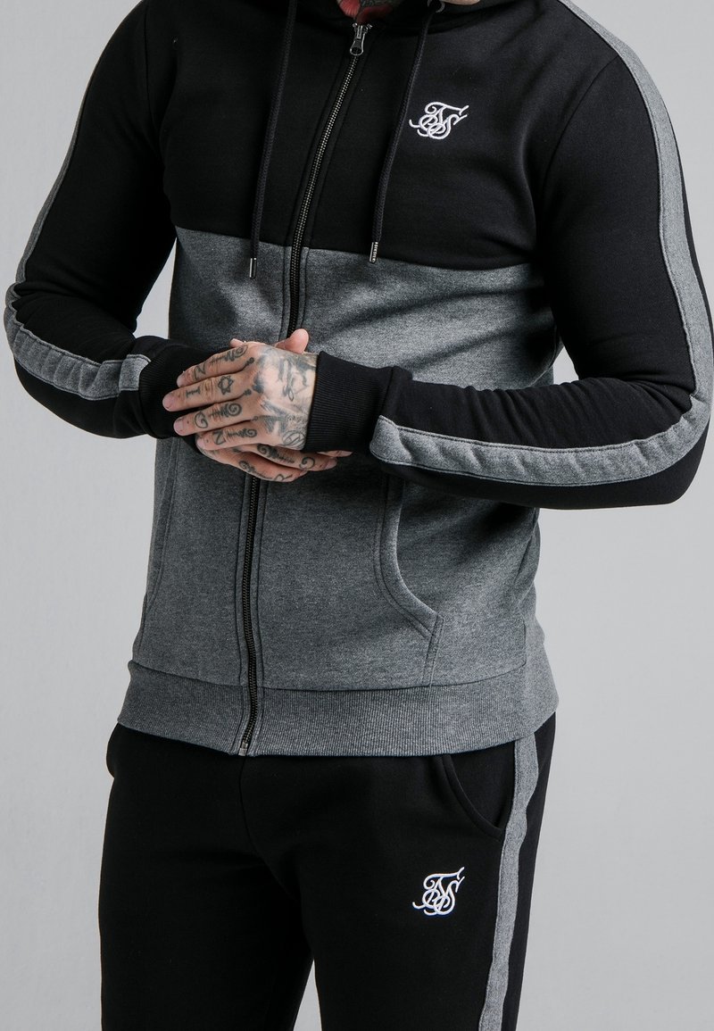 Homme portant un sweat à capuche zippé noir et gris et un pantalon avec des bandes assorties, les mains jointes montrant des tatouages détaillés.