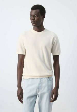 Homme portant un pull en maille crème à manches courtes et un jean bleu clair, debout devant un fond clair uni.