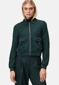 Veste cropped vert foncé en tissu texturé, avec une fermeture éclair sur le devant, un col et deux poches plaquées sur la poitrine. Poignets et ourlet côtelés.