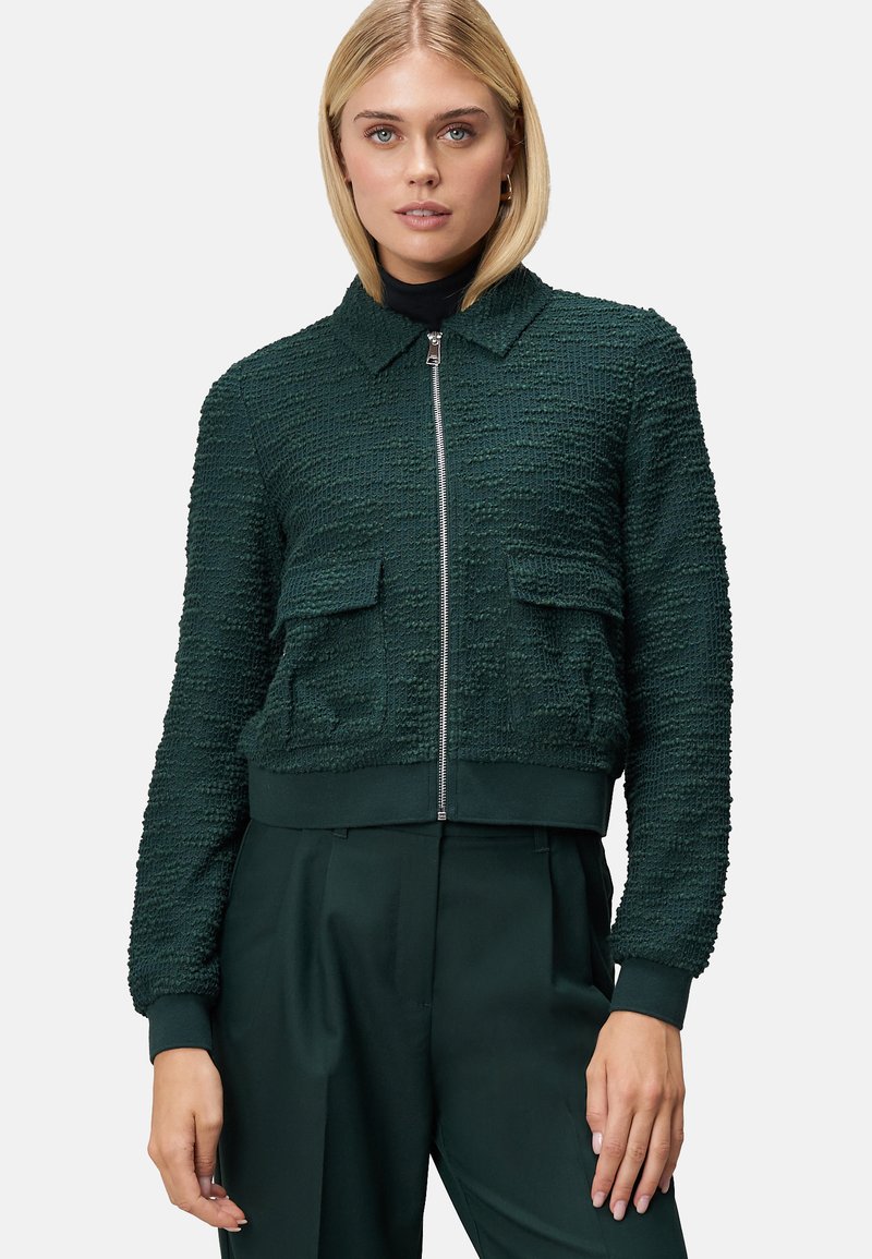 Veste cropped vert foncé en tissu texturé, avec une fermeture éclair sur le devant, un col et deux poches plaquées sur la poitrine. Poignets et ourlet côtelés.