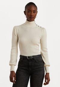 Ribota krēms turtlenecks džemperis ar pūkainām piedurknēm, sānu pogu akcentiem un pieguļošu dizainu, apvienots ar melnām džinsām un melnu jostu.