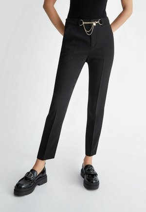 Vrouw draagt zwarte slim-fit broek met een gouden kettingriem en zwarte lakleren loafers op een witte achtergrond.