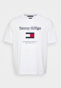 White cotton T-shirt with round neck, featuring "Tommy Hilfiger" logo and a flag emblem in red, white, and blue, with established date below.