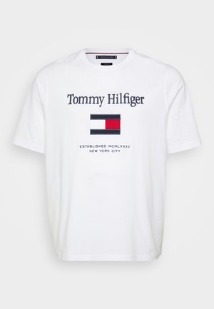 Vit bomulls-T-shirt med rund hals, med "Tommy Hilfiger"-logotyp och en flaggemblem i rött, vitt och blått, med etableringsdatum nedanför.