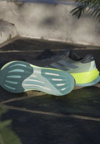 Atletische schoenen met een grijze bovenkant, een mesh-structuur en sprankelende gele accenten. De rubberen zool heeft een golvend patroon in teal en lichtblauw.
