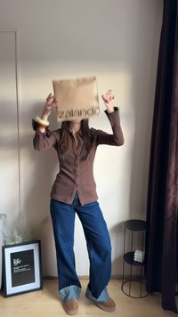 Brown button-up shirt, wide blue denim jeans with frayed hems, and tan footwear. Holding a brown paper bag with "zalando" logo.