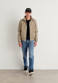Lys beige bomberjakke med en svart t-skjorte, blå jeans og svarte sneakers. Jakken har stående krage og mansjetter.