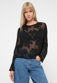 Sort gennemsigtig bluse med blomster applikation, lange ærmer, rund halsudskæring, parret med lyse denim jeans. Blød tekstur med lagdelte detaljer.