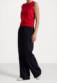 Top en tricot sans manches rouge associé à un pantalon large bleu marine avec des poches latérales et des accents de boutons, porté avec des chaussures de couleur claire.