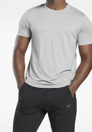 Homme portant un t-shirt à manches courtes gris clair et un pantalon de sport noir, mains dans les poches, sur fond blanc.