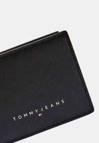 Cartera de cuero negra con el logotipo en relieve "TOMMY JEANS" en blanco. Presenta líneas limpias, costuras visibles y un acento de color debajo del logotipo.