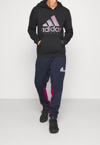 Svart hoodie med lila gradient Adidas-logotyp, framficka; marinblå joggers med rosa sidopaneler och vit Adidas-logotyp på låret.