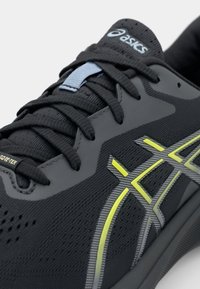 Svart sportskor med en strukturerad ovandel, grå och gula accenter samt en slät, dämpad sula. Har en snörningsdesign och logotyp.