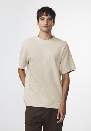 Beige strukturiertes Kurzarm-T-Shirt mit Rundhalsausschnitt, locker sitzend und mit subtiler Markenkennung nahe dem Saum. Getragen mit dunkelbraunen Hosen.