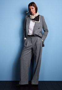 Gray tailored suit with a double-breasted jacket, pleated wide-leg trousers, and a white shirt. Accompanied by a black and white polka dot scarf.