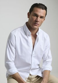 Camisa blanca de botones de manga larga con cuello clásico y puños enrollados. Tejido suave con un sutil detalle de logo en el pecho.