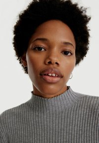 Grauer gerippter Pullover mit hohem, gezacktem Ausschnitt. Das Model hat natürliche Haare und trägt kleine Creolen, vor einem neutralen Hintergrund.