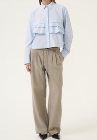 Lichtblauw shirt met frontale ruches, gecombineerd met beige geplooide broeken met een trekkoord in de taille; zwarte puntige schoenen.