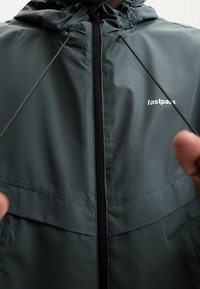 Zbliżenie osoby w ciemnoszarej kurtce na zamek z kapturem, ściągającej czarne sznurki, z białym logo "fastpass" na piersi.