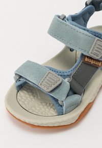 Ljust blå mockaskor med grå detaljer och justerbara remmar. Texturerad grå sula med en brun gummibotten för grepp.