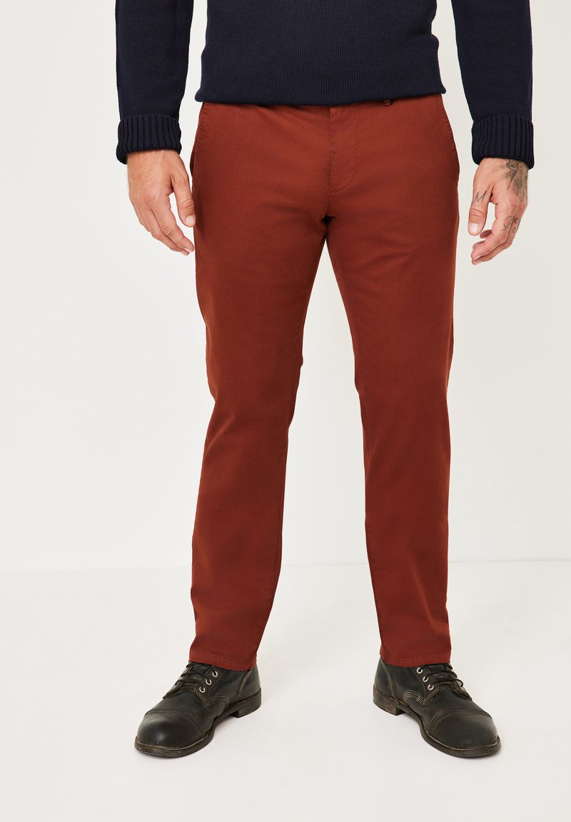 Rust-colored cotton trousers with a slim fit, featuring pockets and stitching details, paired with black lace-up shoes.