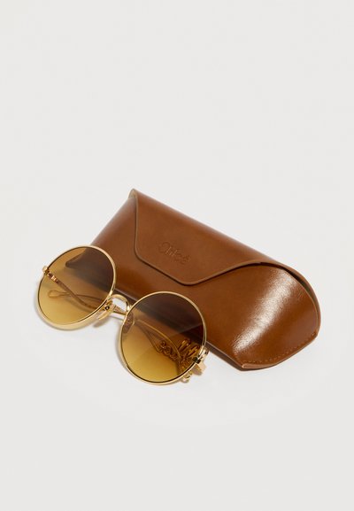 Στρογγυλά γυαλιά ηλίου με χρυσό πλαίσιο και καφέ διαβάθμιση, που ακουμπούν σε καφέ δερμάτινη θήκη με το ανάγλυφο λογότυπο "Chloé".