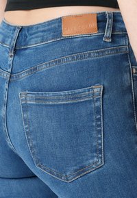 Plave traper hlače s visokim strukom, s laganim šavovima, stražnjim džepovima i svijetlosmeđom kožnom etiketom s natpisom "BLUE DENIM" urezanim na njoj.