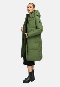 Kvinna klädd i en knälång olivgrön vadderad kappa med huva, svart klänning och svarta klumpiga stövlar, som står på sidan mot en enfärgad bakgrund.