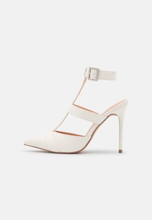 Zapato de tacón stiletto blanco de punta puntiaguda con correa en el tobillo y hebilla plateada, con dos correas horizontales sobre el pie.