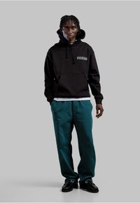 Felpa con cappuccio nera con tasca frontale, dettagli in bianco. Abbinata a pantaloni larghi color teal e scarpe nere.