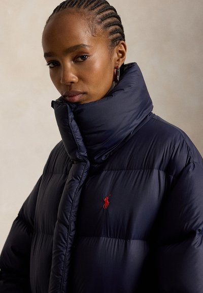 Femme aux cheveux tressés porte une doudoune bleu marine avec un col montant et un logo rouge brodé sur la poitrine, faisant face vers l'avant.