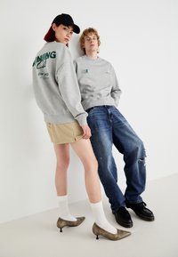 Grijze sweatshirt, één met groene grafische tekst; beige shorts en verslete blauwe jeans; gouden hakken; witte sokken; minimalistische achtergrond.