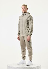 Light beige lightweight jacket with a hood and elastic cuffs, paired with matching beige tapered pants and white sneakers. Smooth textures.