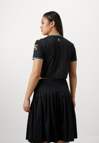 Crni skraćeni džemper s kratkim rukavima sadrži plave i žute akcentne boje na ovratniku i rukavima, u kombinaciji s plisiranom crnom suknjom.