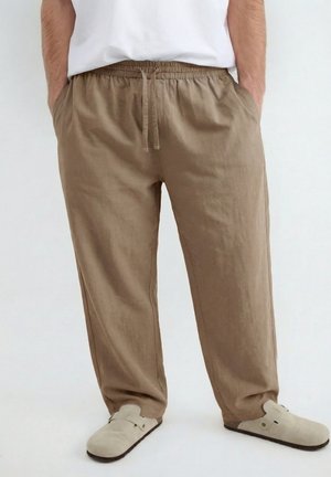 Man draagt losse beige broek met trekkoord, handen in zakken en lichtbeige slip-on schoenen op een effen achtergrond.