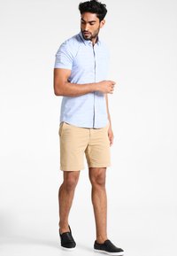 Chemise à manches courtes bleu clair à rayures, associée à un short beige et des chaussures noires à enfiler. Tenue d'été simple et décontractée.