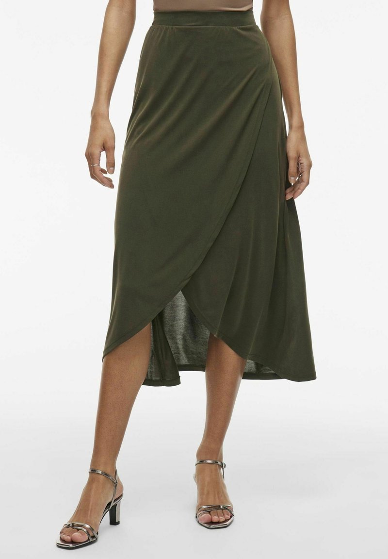 Femme portant une jupe midi portefeuille vert olive et des sandales à talons métalliques, debout contre un fond clair uni.