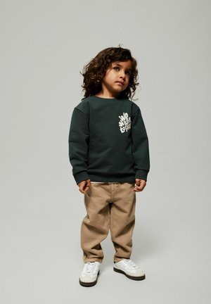 Ciemnozielona bluza z białym nadrukiem, beżowe luźne spodnie i białe trampki. Dziecko ma kręcone ciemne włosy. Prosty, codzienny strój.